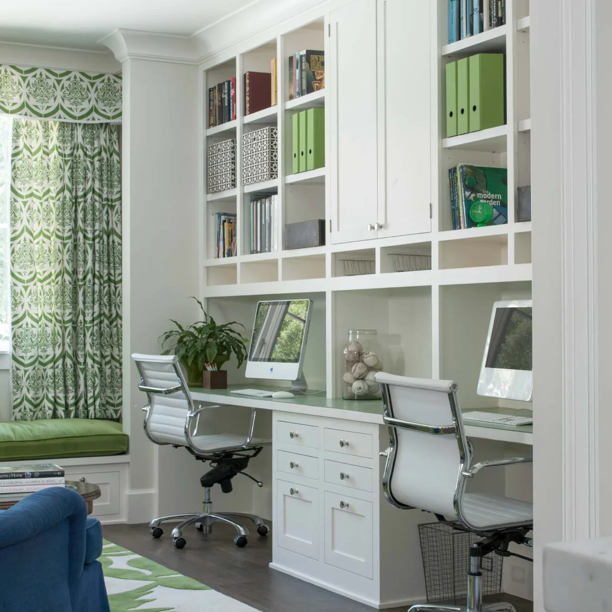 Two computer workstations in white fitted cabinetry beside a blue sofa and green window-seat cushion.