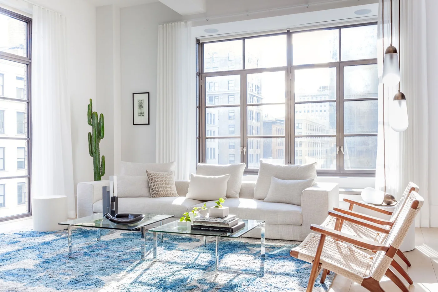 A white sofa and two open-framed wood chairs face glass tables on a blue patterned rug beside broad apartment windows