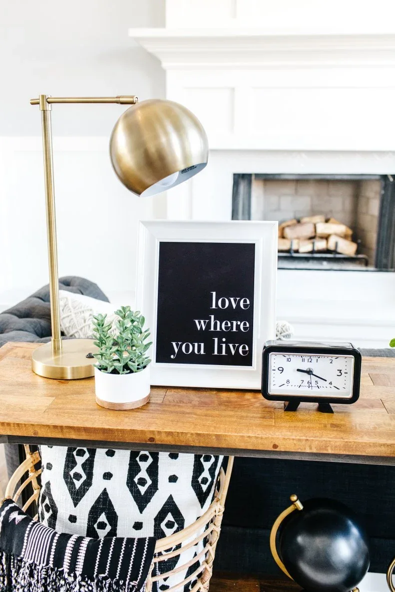 A slim gold-toned lamp sits beside a framed print, small plant and clock on a timber sofa table.