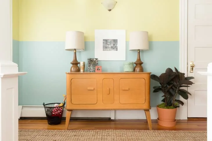 A smaller orange sideboard with rounded rectangular front details stands beneath two lamps against a yellow-and-blue wall beside a potted plant