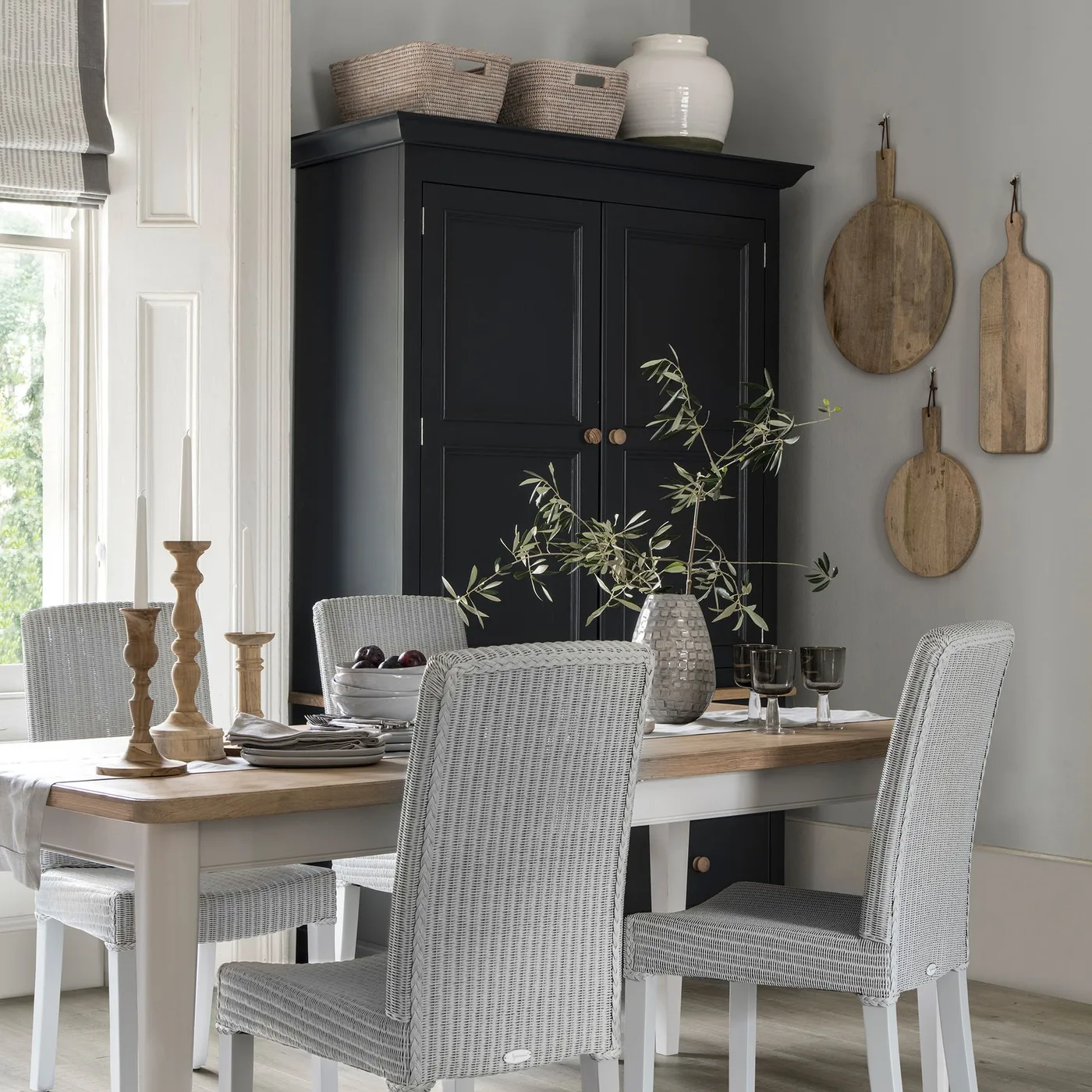 Country dining room with black paneled cupboard, pale table, woven light chairs, wooden cutting boards and baskets above the storage.