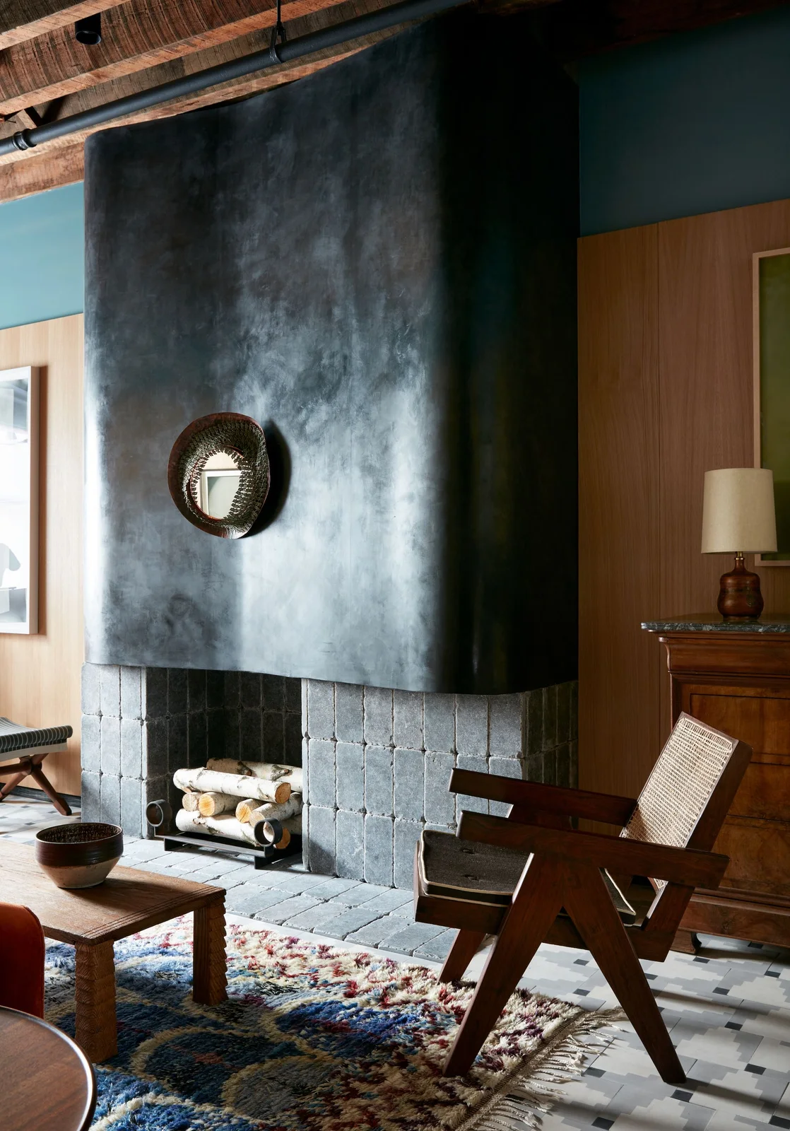 A tall dark curved fireplace hood rises above gray hearth tiles beside a wood-framed chair, wood wall panels and a small round mirror