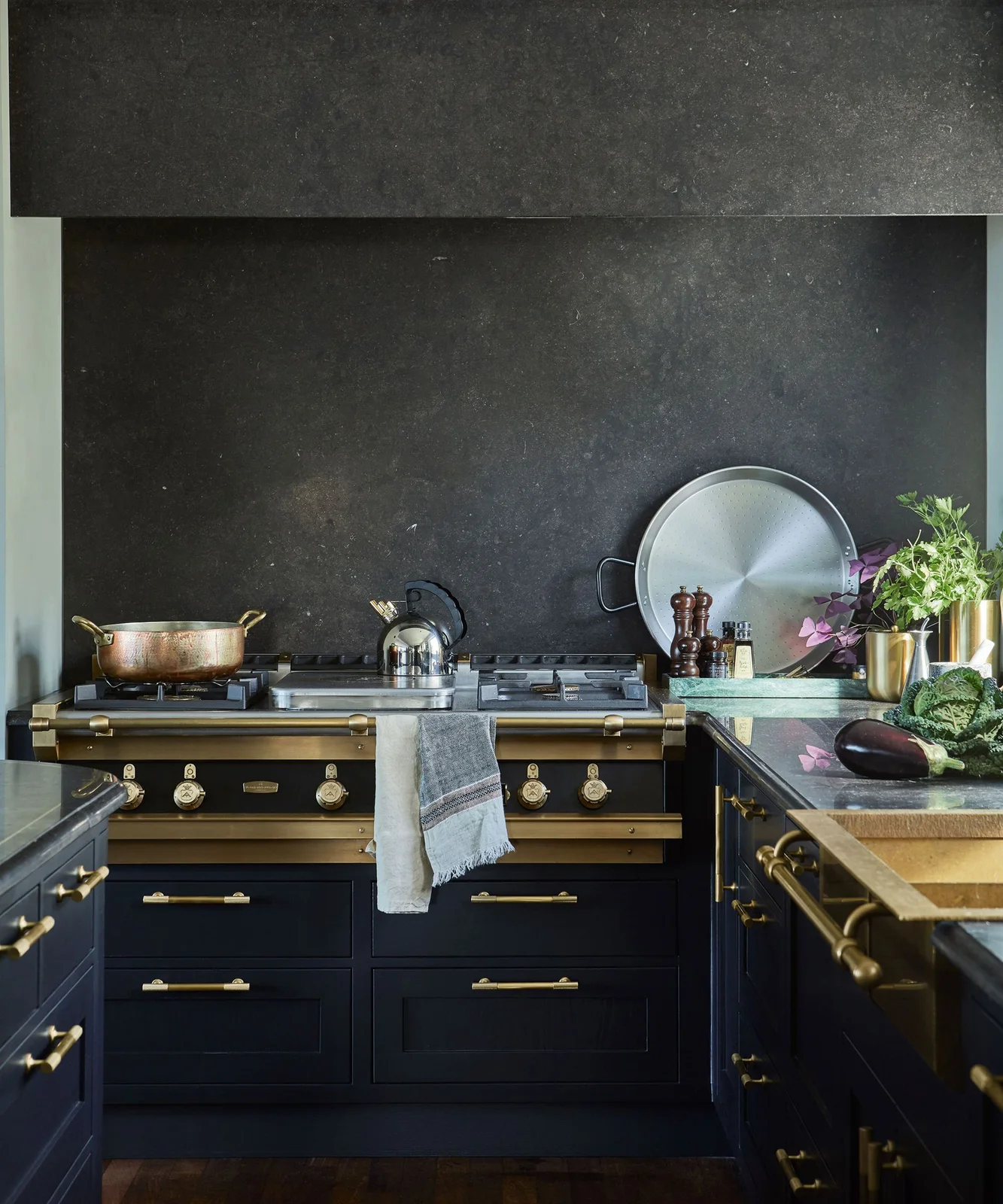 A dark cooker with warm-colored knobs sits among navy cabinets below a dark hood beside a green-toned counter, cookware and sink