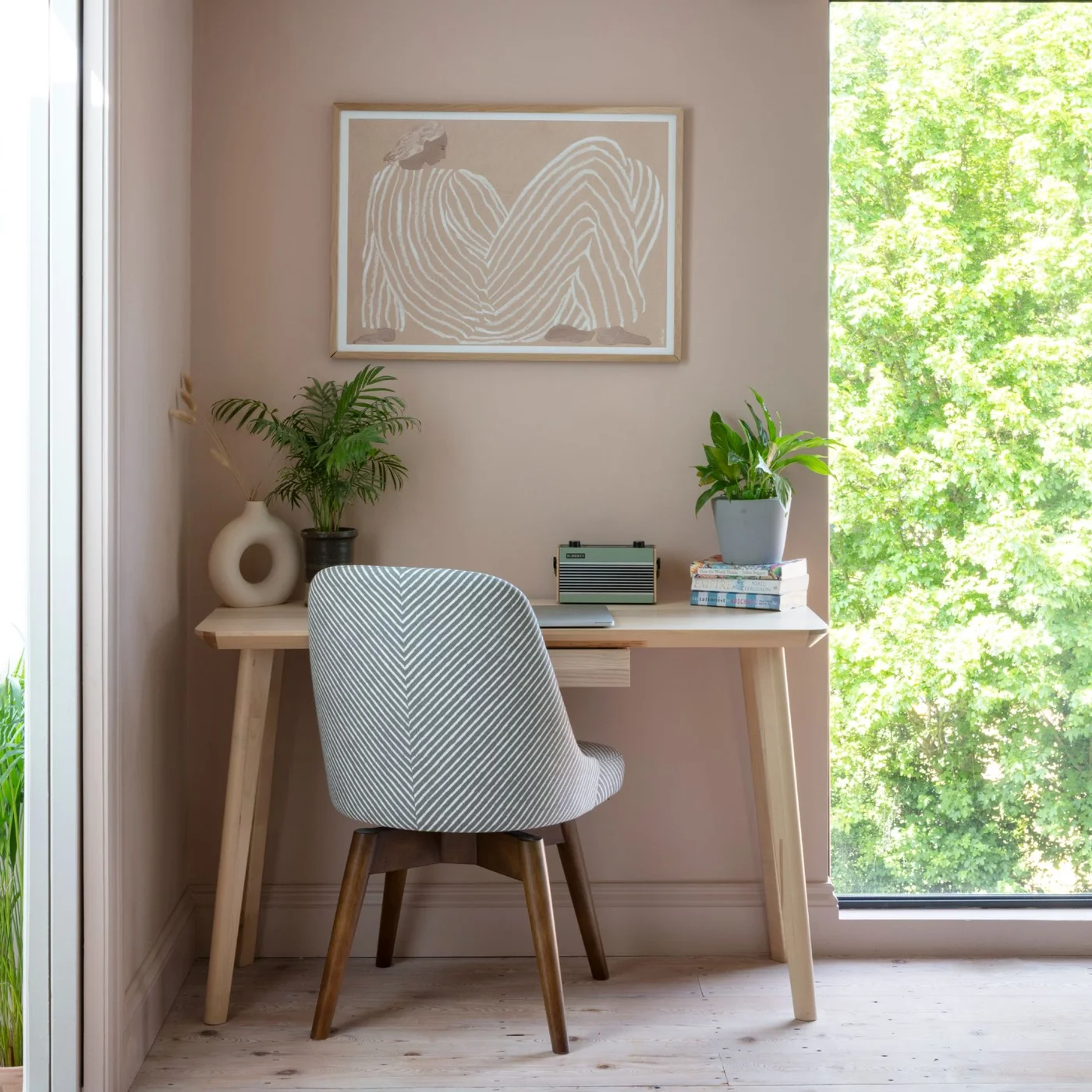A small pale wood desk and gray patterned chair sit against a blush wall beside tall glazing, with a tonal framed print and plants at the desk edges.