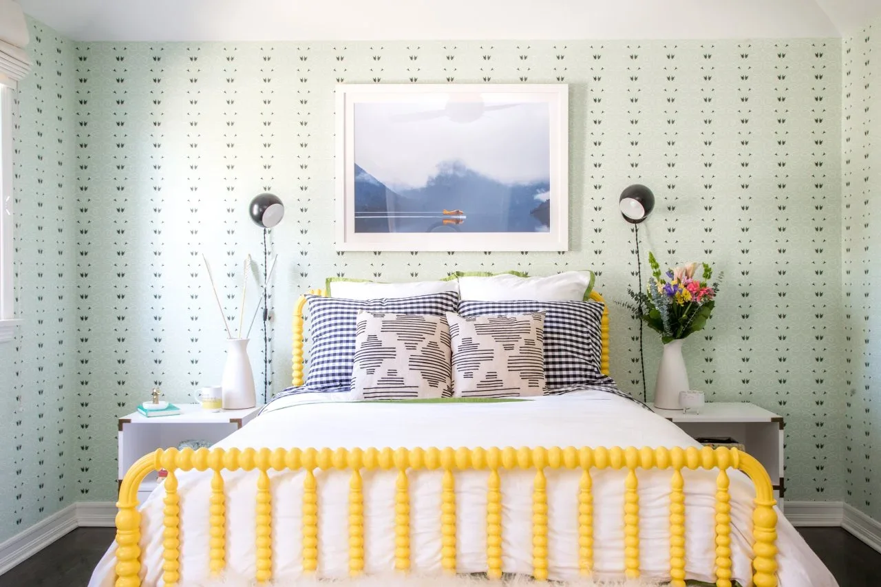 A bright yellow spindle bed has black-and-white checked pillows against a pale mint patterned wall beneath a large blue landscape picture.
