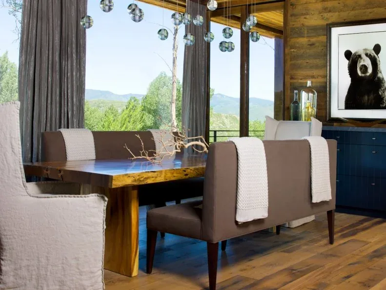 Dining room with horizontal brown wood wall behind bear artwork and dark blue cabinetry, wooden table, upholstered seating, glass globe lights, and broad windows.