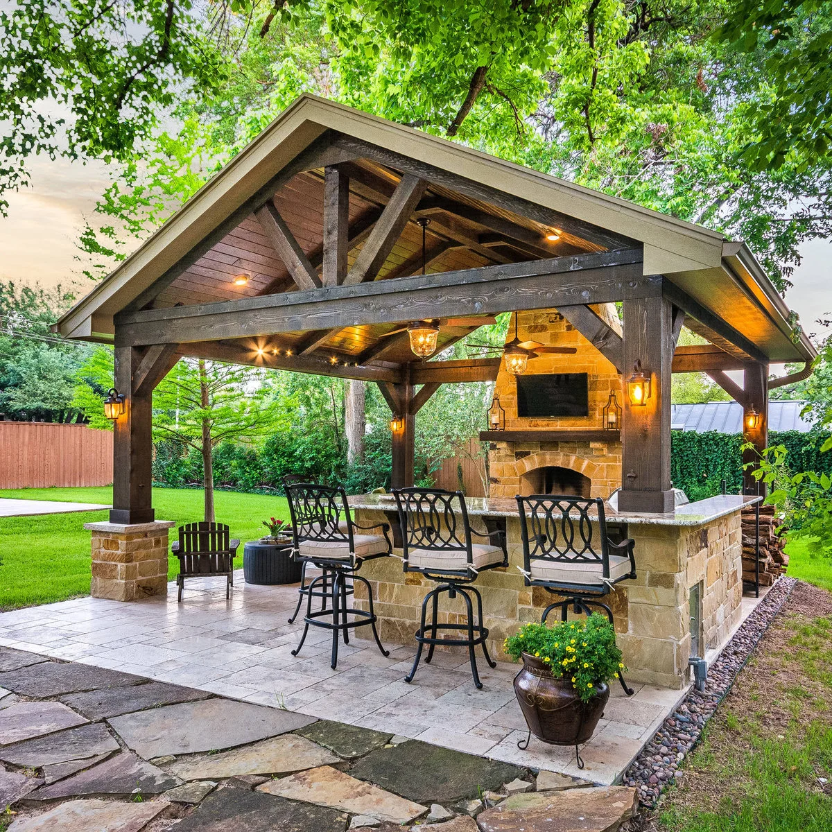 Stone bar with three ornate dark stools beneath a timber pavilion with a fireplace and garden path approaching