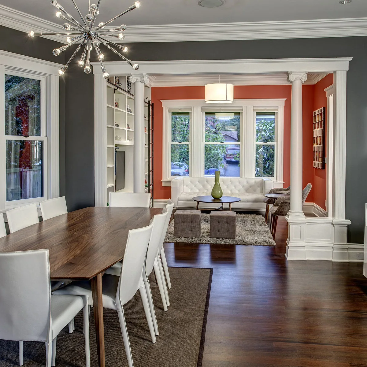 Dining room with a wooden table and white chairs opening between columns into a small orange-walled sitting room with a white sofa.