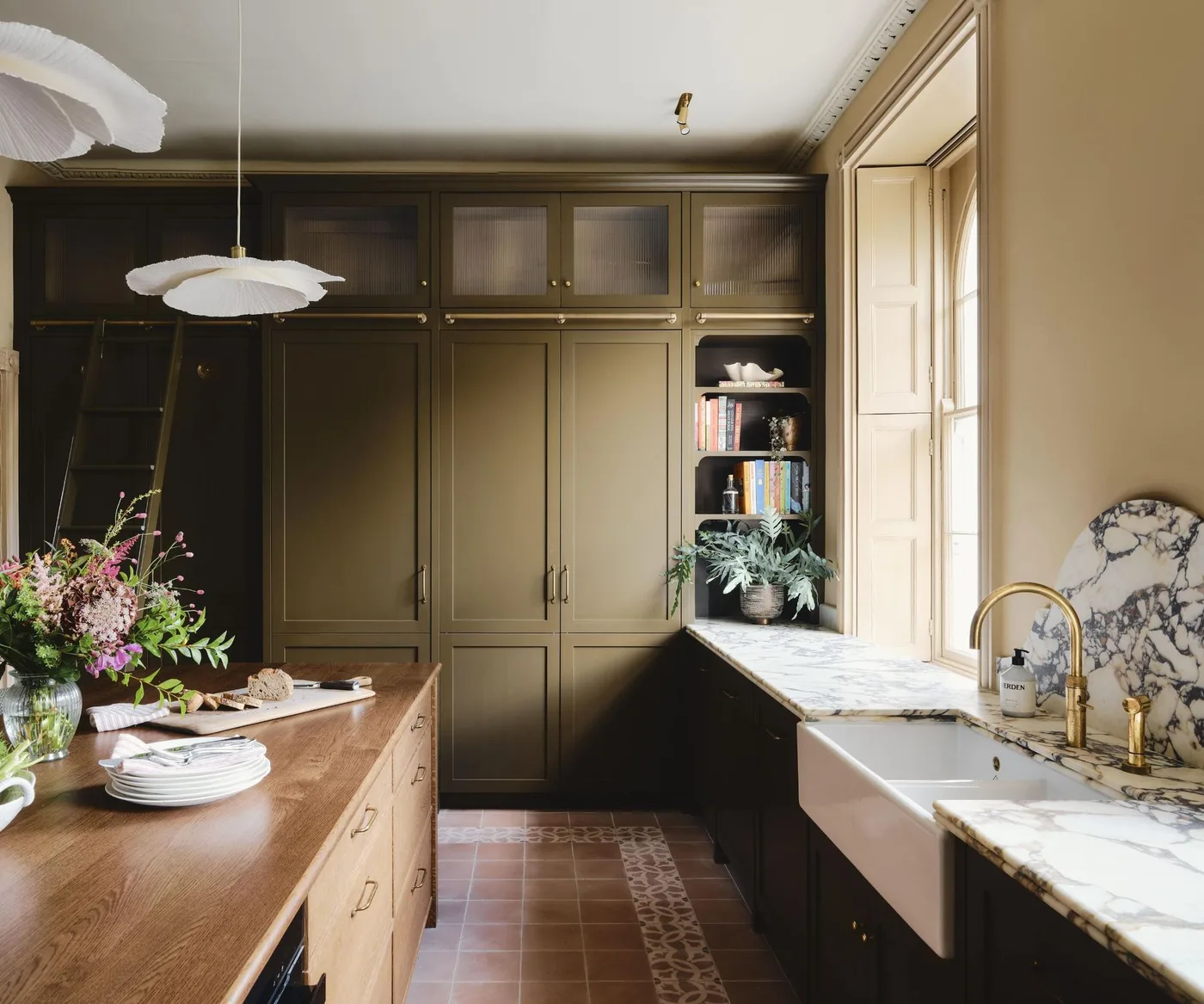 Dark olive kitchen cabinets with a white apron-front sink and a pale veined backsplash rising in a curve beneath the window.