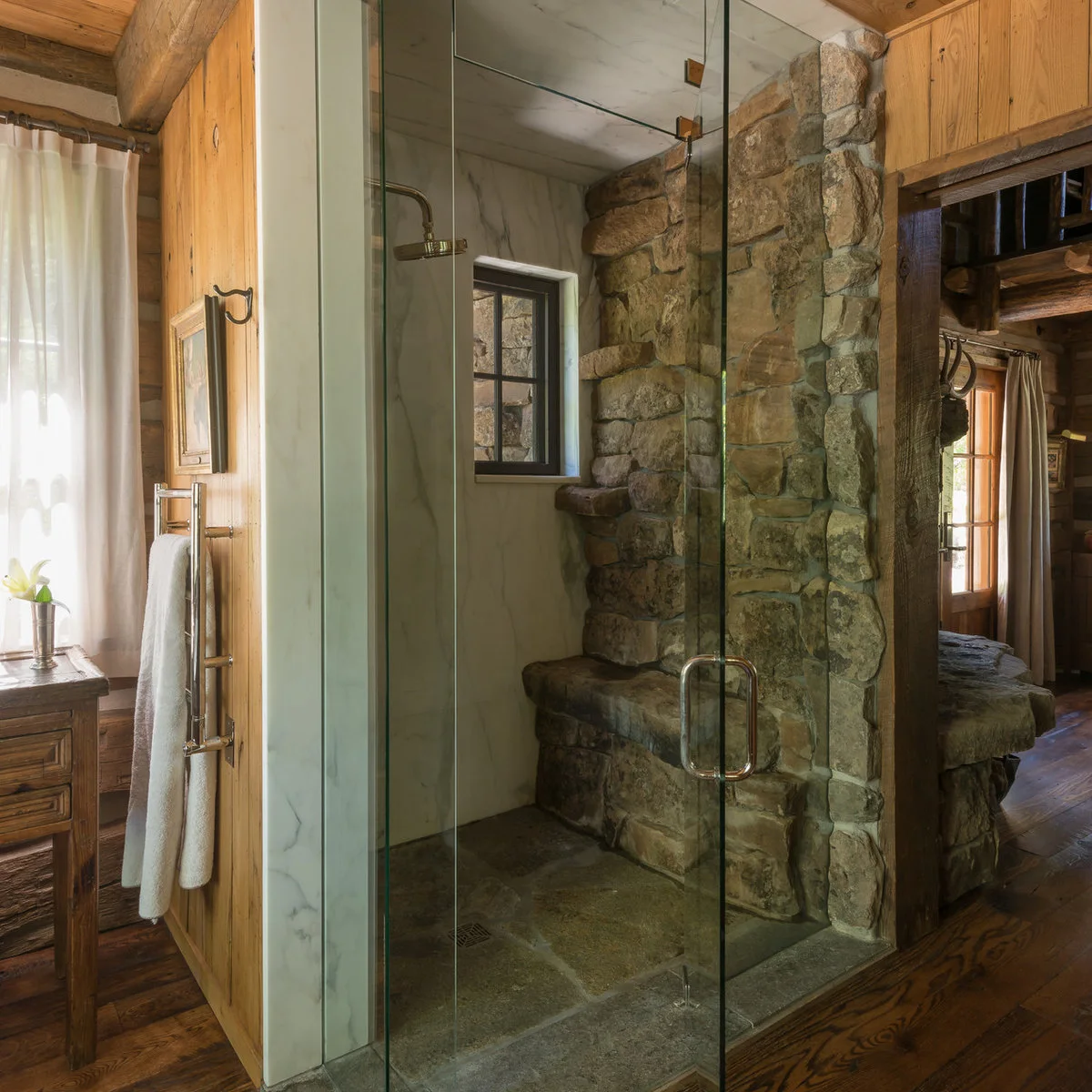 A glass shower with one rough stone-textured wall, a built-in seat and pale veined surfaces on the neighboring wall and ceiling.
