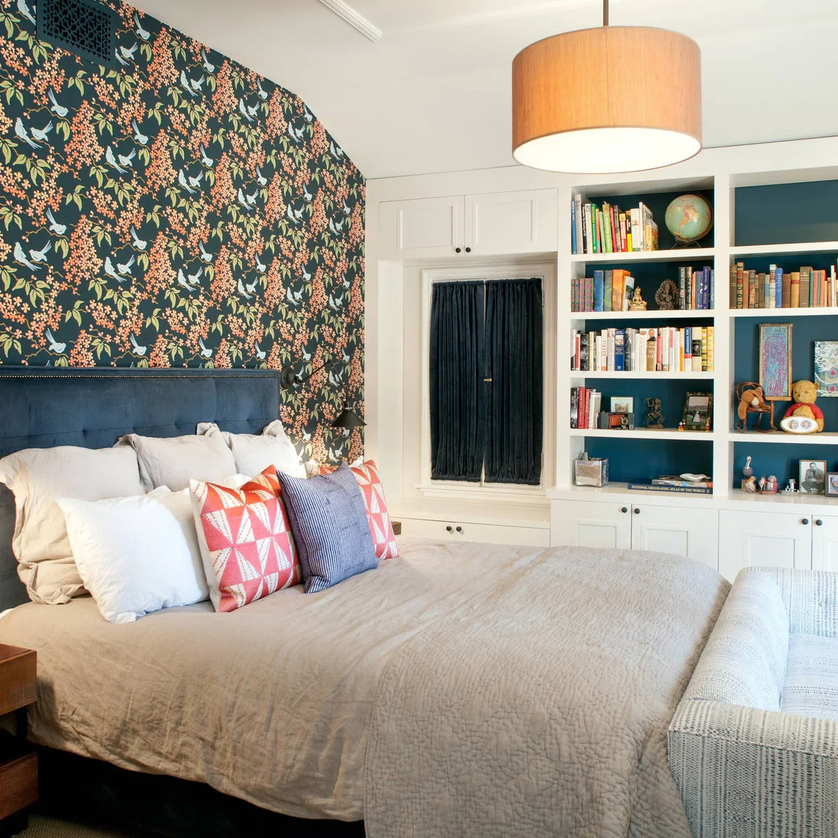 Dark bird and flowering-branch wallpaper behind a blue headboard beside white bookshelves with blue backs.