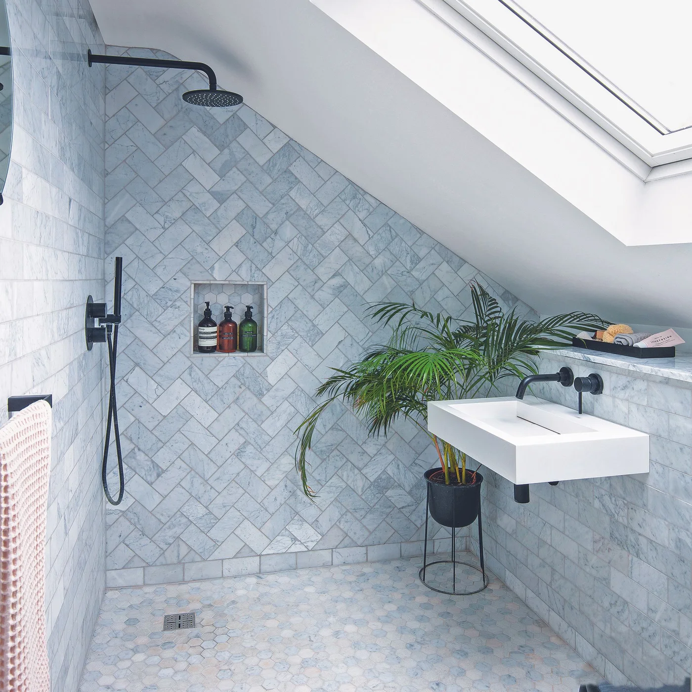 An attic shower room has gray herringbone wall tiles, small hexagonal floor tiles, a rooflight, black shower fittings and a compact white wall-mounted basin.