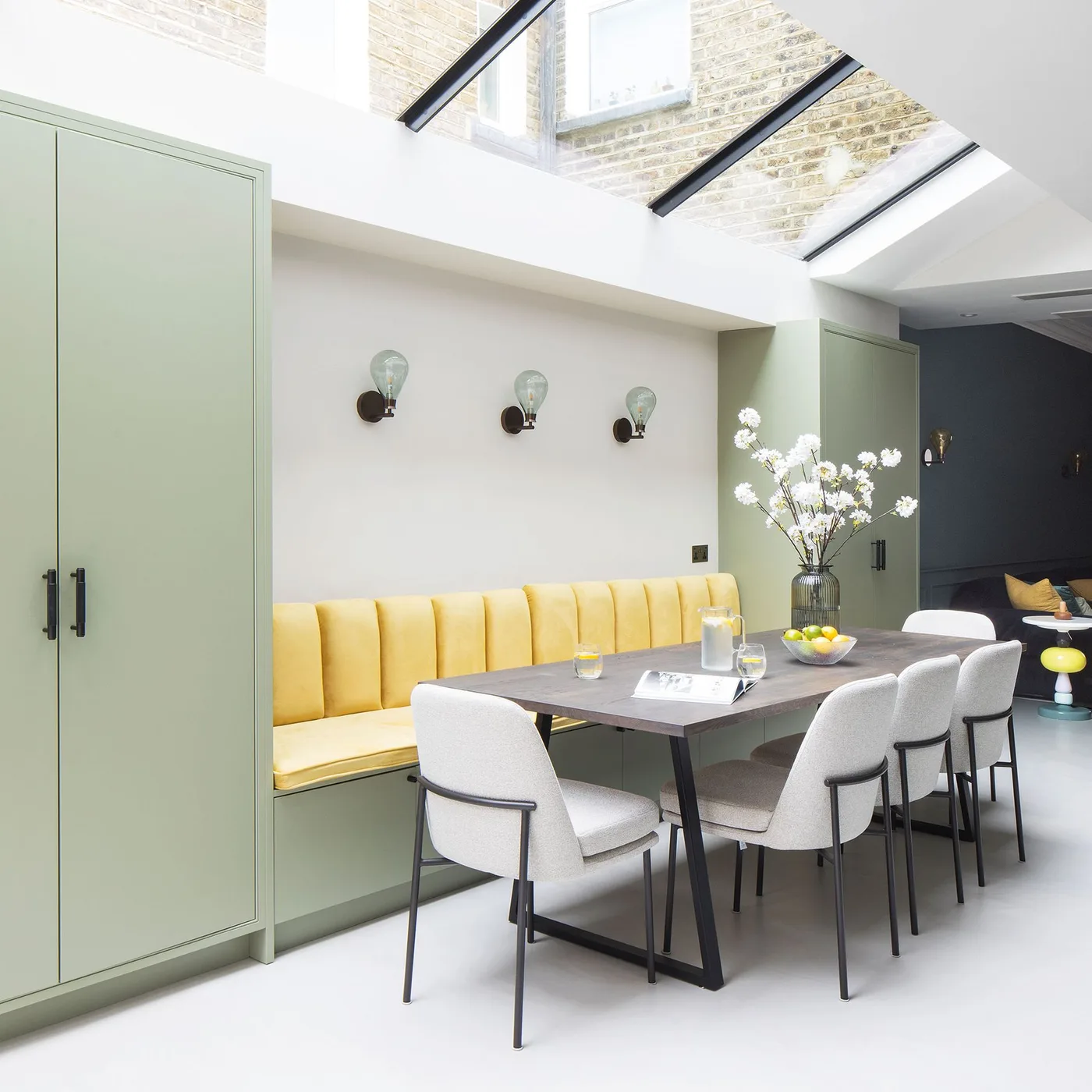 Dining space with a yellow upholstered banquette, dark rectangular table, pale upholstered chairs, green cabinets and roof windows.