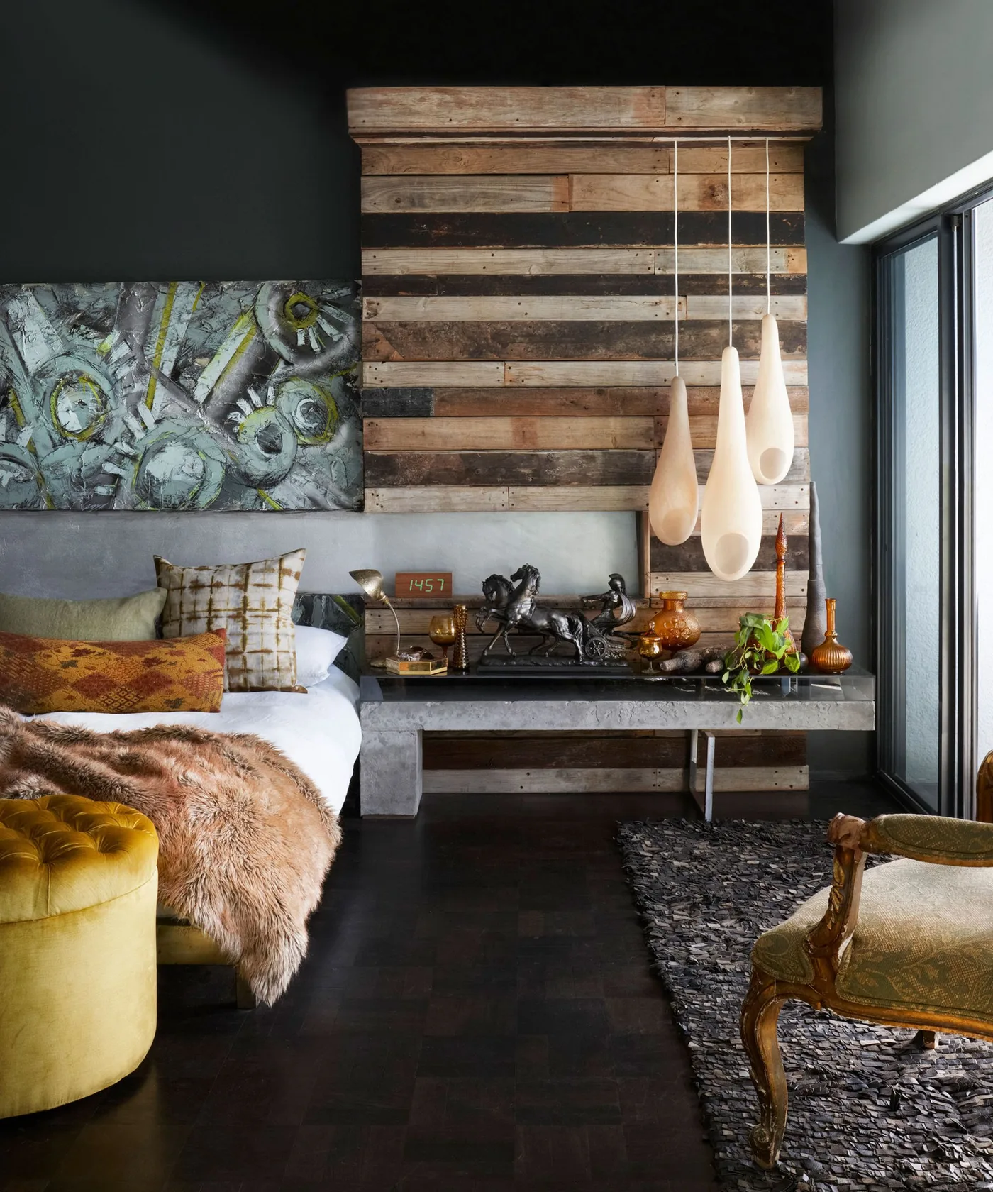 Dark bedroom with a wood-plank wall section, pale hanging lights, patterned cushions, shaggy throw, and artwork beside the bed.
