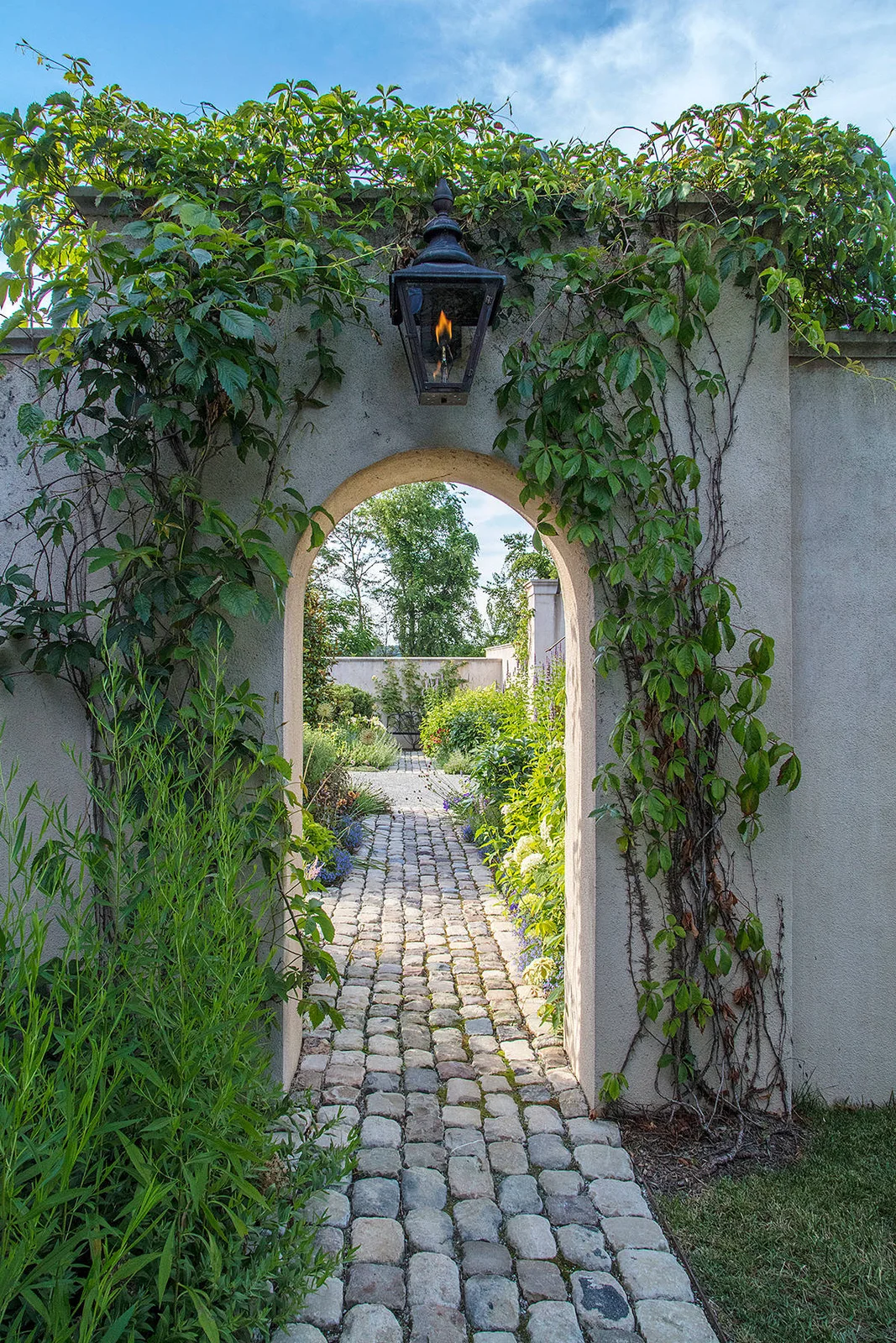 A narrow cobbled path passes through a vine-covered arch toward a garden table