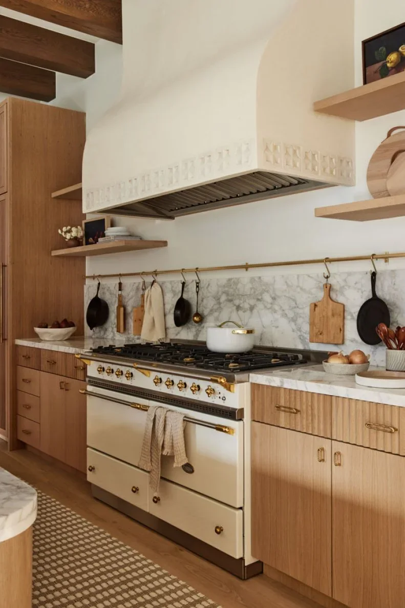 Fluted wood drawer fronts and smooth lower cabinet doors sit beside a cream range under a pale hood and wood shelves.