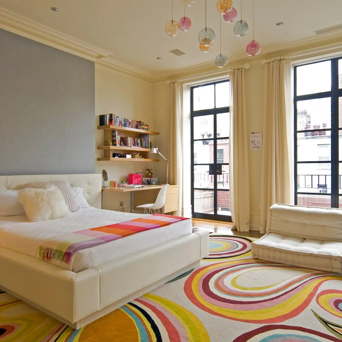 Teen bedroom with a desk and shelves beside tall windows, white furniture and a colorful curved-pattern rug