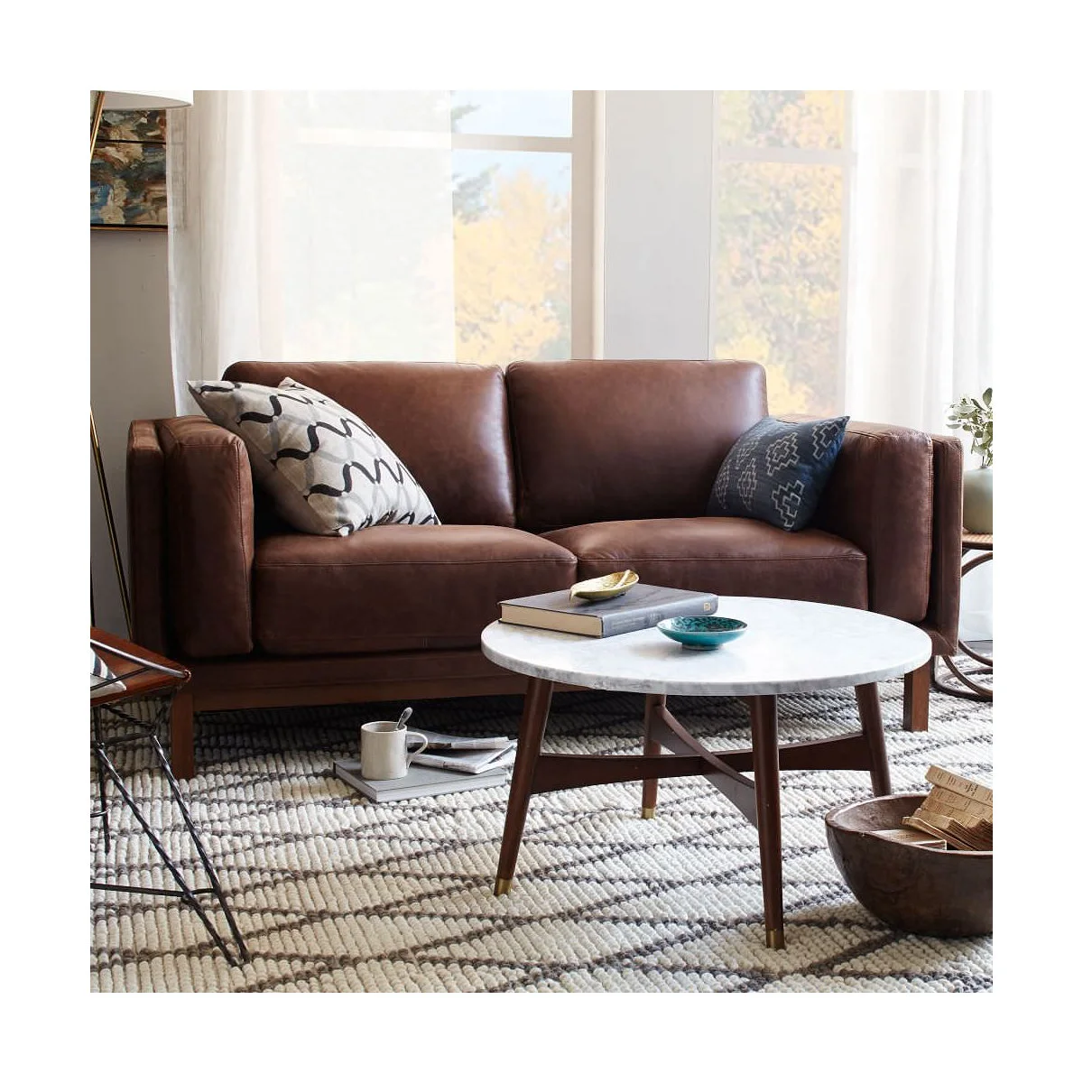 Brown leather two-seat sofa with pale cushions beside a round white coffee table and gray rug.