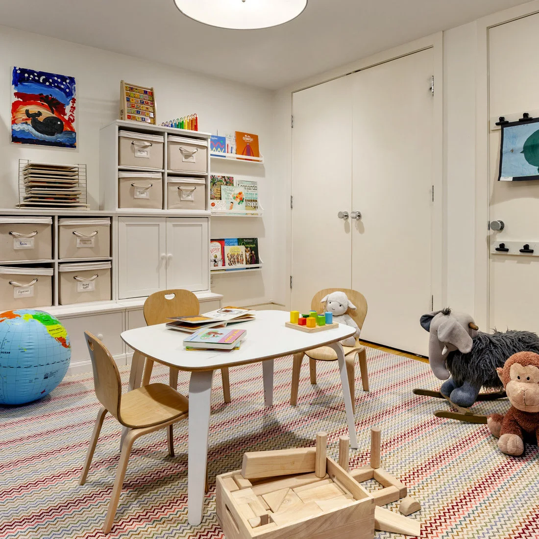 A white playroom has a small white table with wood legs, wood chairs, labeled fabric bins, face-out books and wooden blocks on a patterned rug.