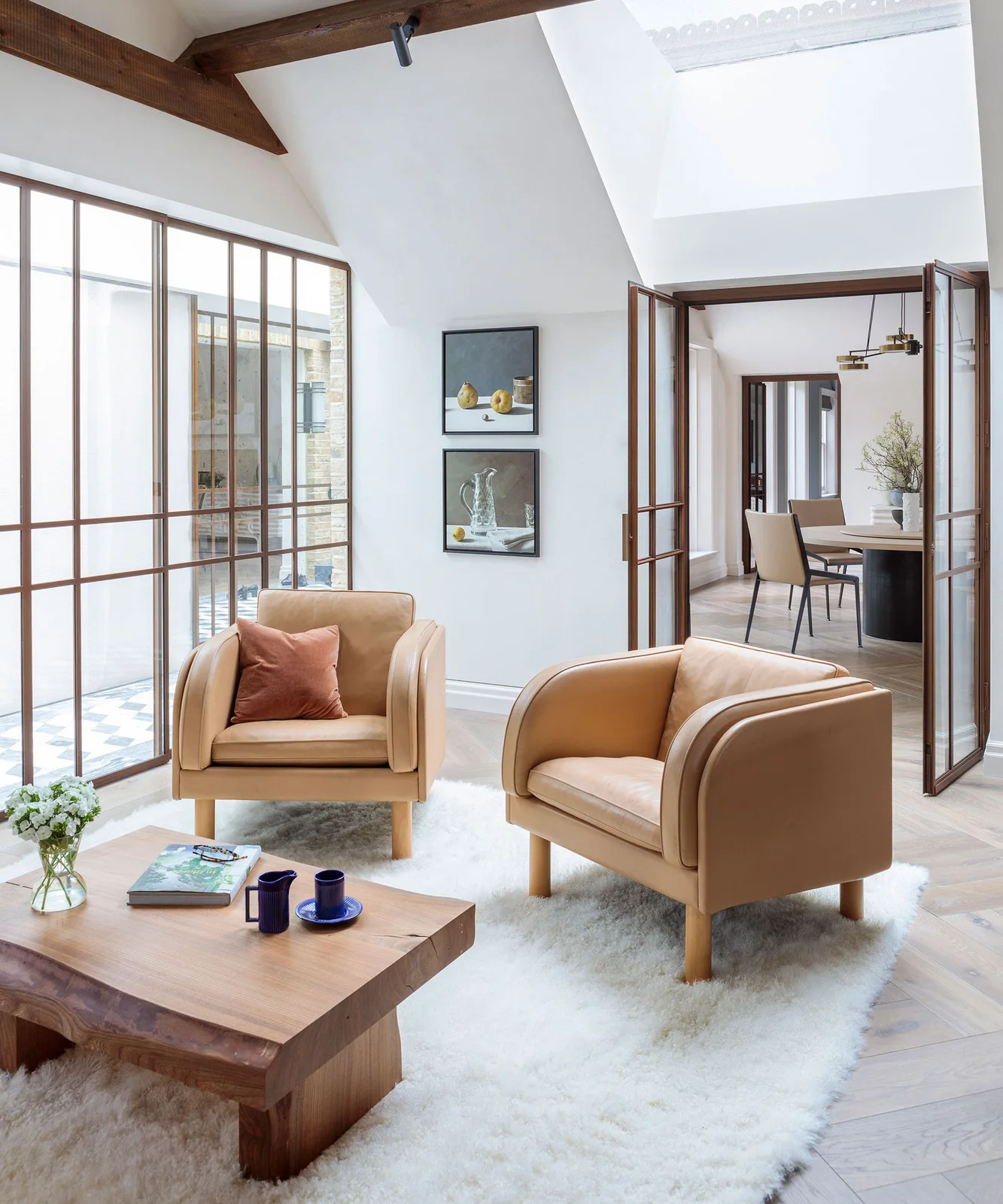 Two tan chairs and a low wood table sit on a pale rug beside wood-framed glazed partitions and a view into a dining area