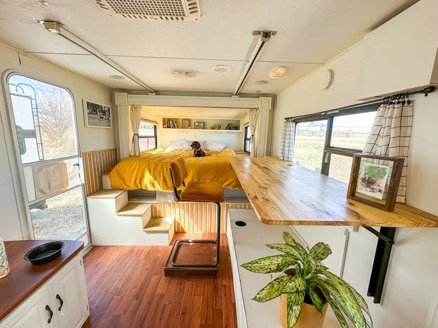 Camper with a long wood work surface beneath a window, an ochre chair and steps leading to a yellow-covered bed.