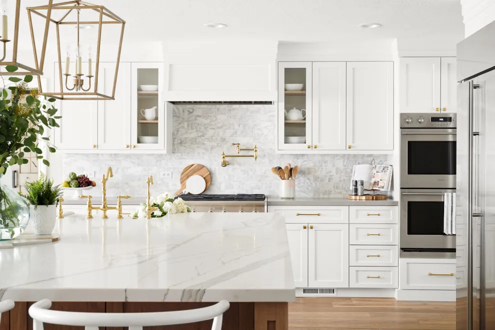 White cabinets and glass-front cupboards surround a pale worktop with warm metal fixtures