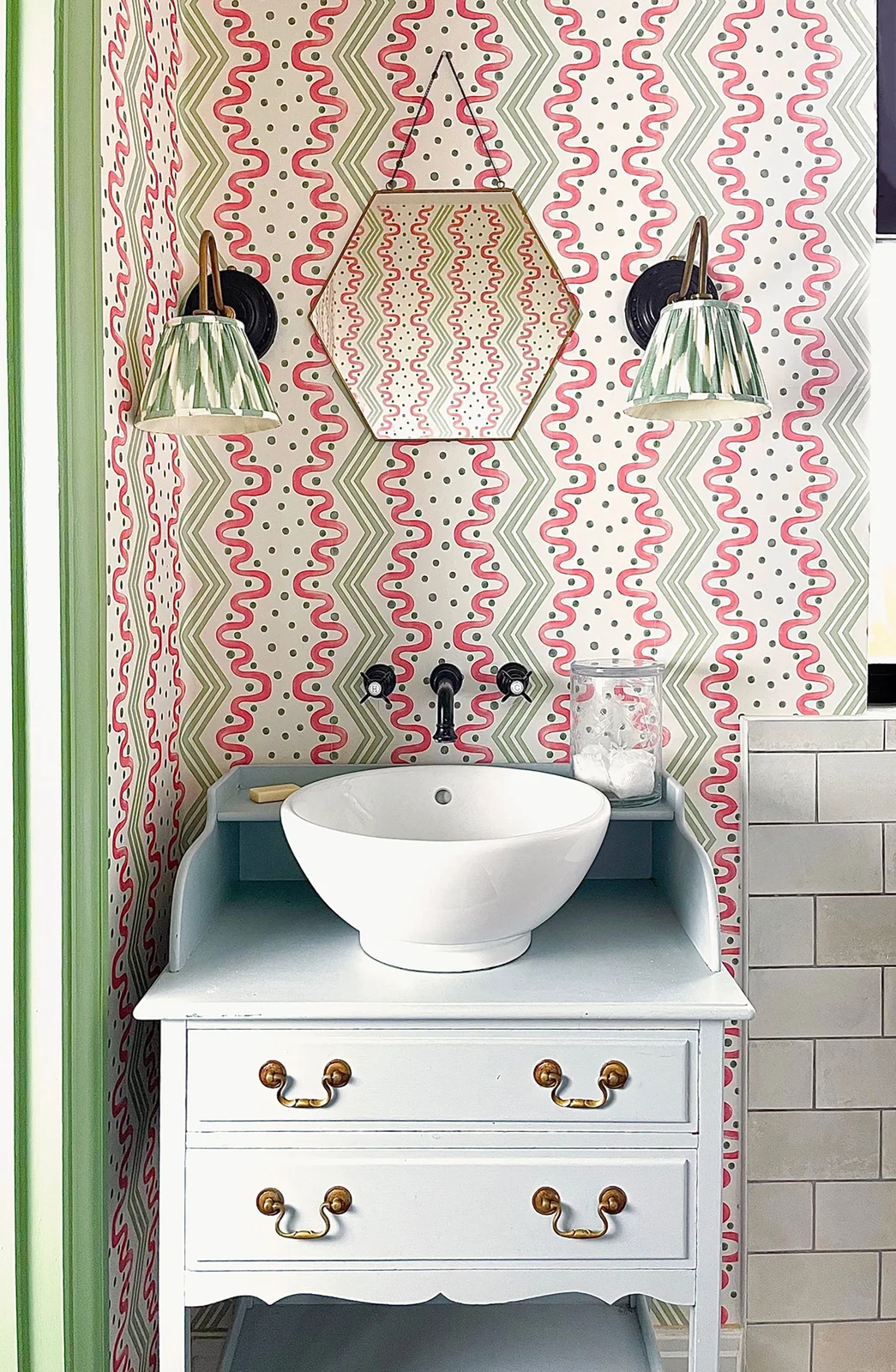 Pink-and-green wavy wallpaper surrounds a pale vanity beneath a hexagonal mirror and green patterned light shades.