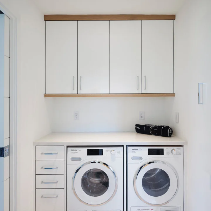 Two front-loading laundry machines sit beneath a white counter and white wall cupboards framed with pale wood.