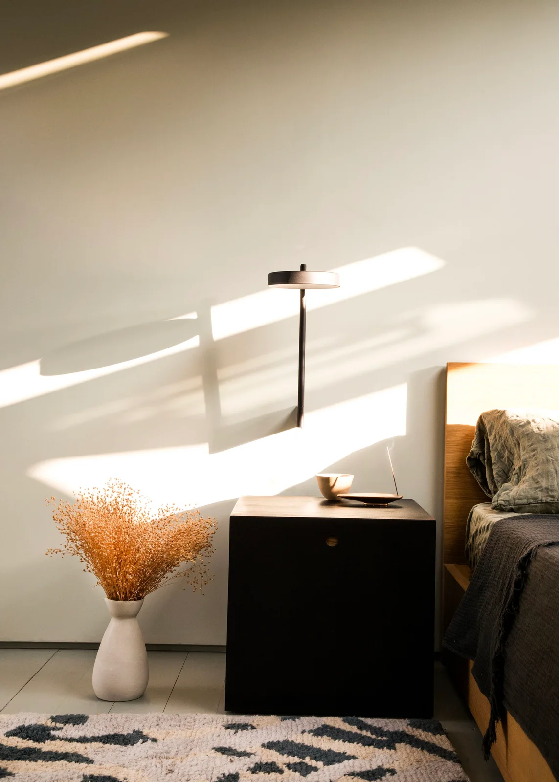 A dark cube-like bedroom cabinet stands beside a wood bed frame beneath a slim projecting wall light and pale wall with a patterned rug below