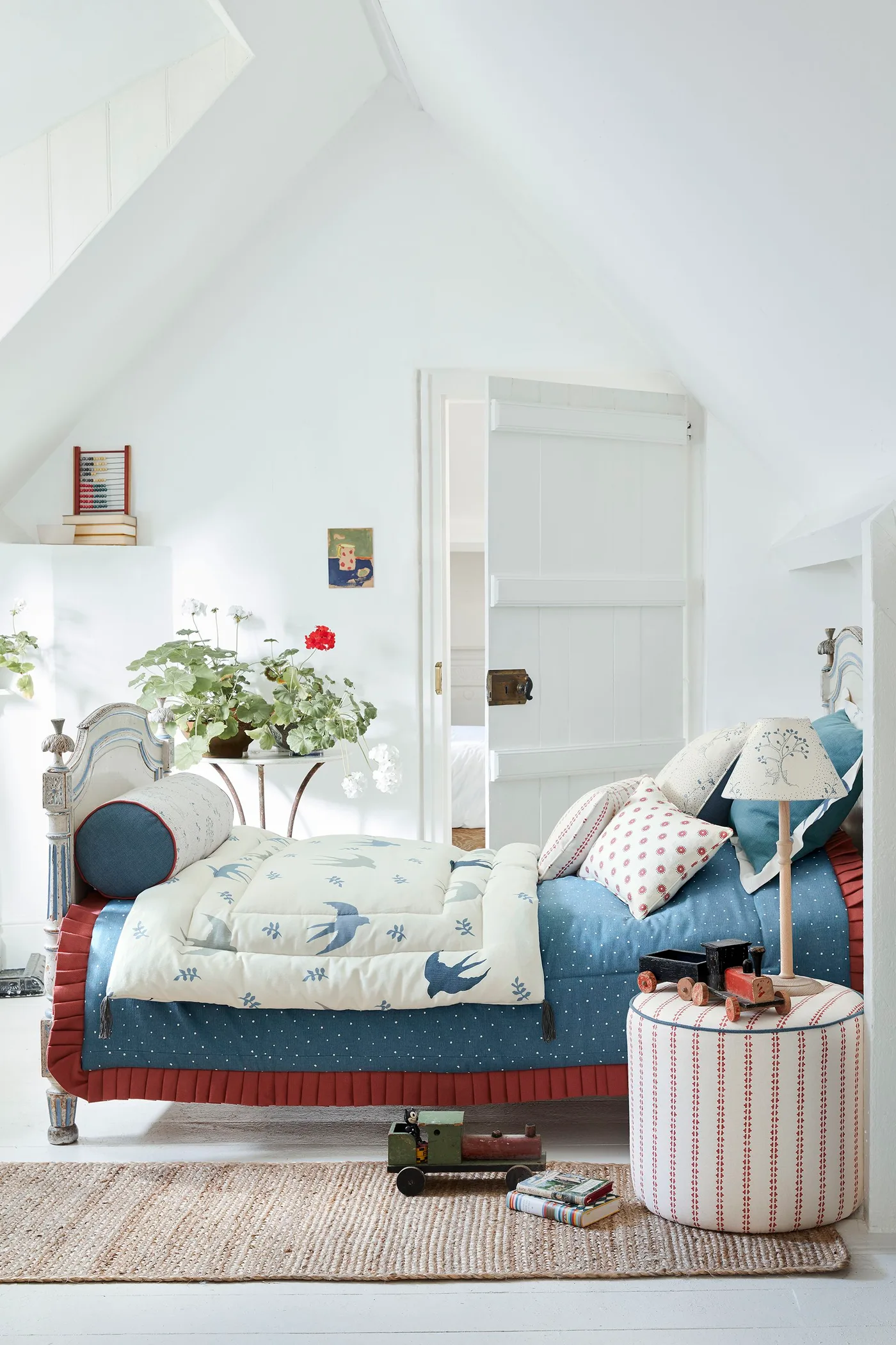 Pale attic bedroom with a blue bedcover, red pleated bed skirt, patterned cushions, striped round table and floral armchair.