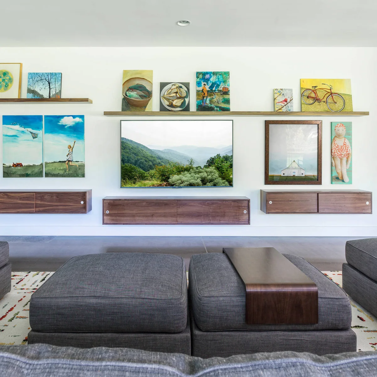 Long wood picture ledge with small colorful paintings above a landscape-displaying screen and floating wood cabinets.