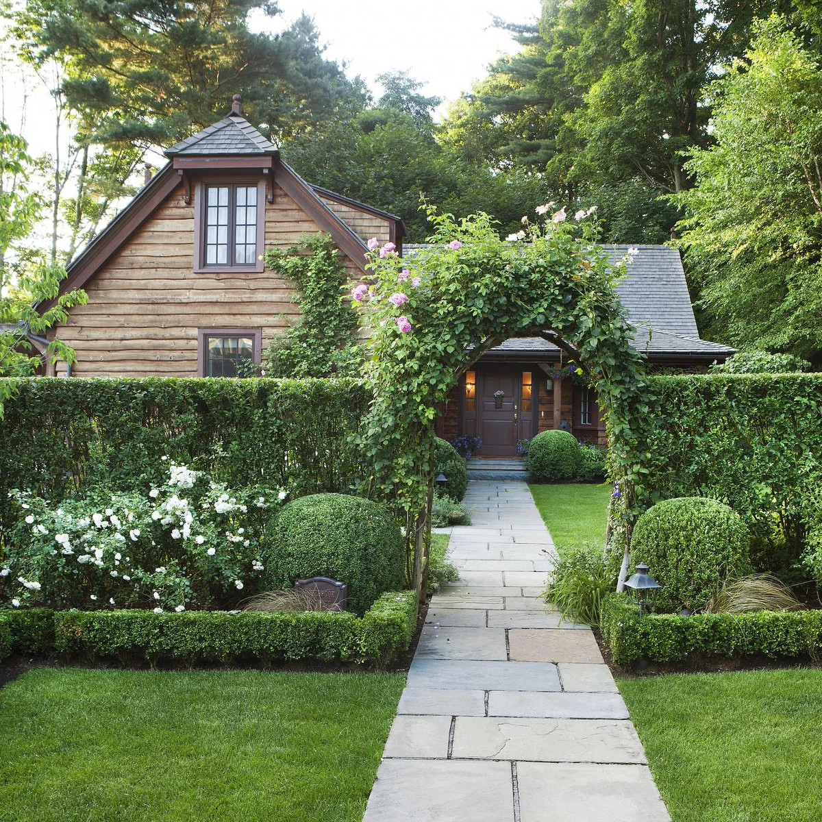 A flower-covered arch aligns with a front door beyond a straight pale path, clipped hedges and a small lawn.