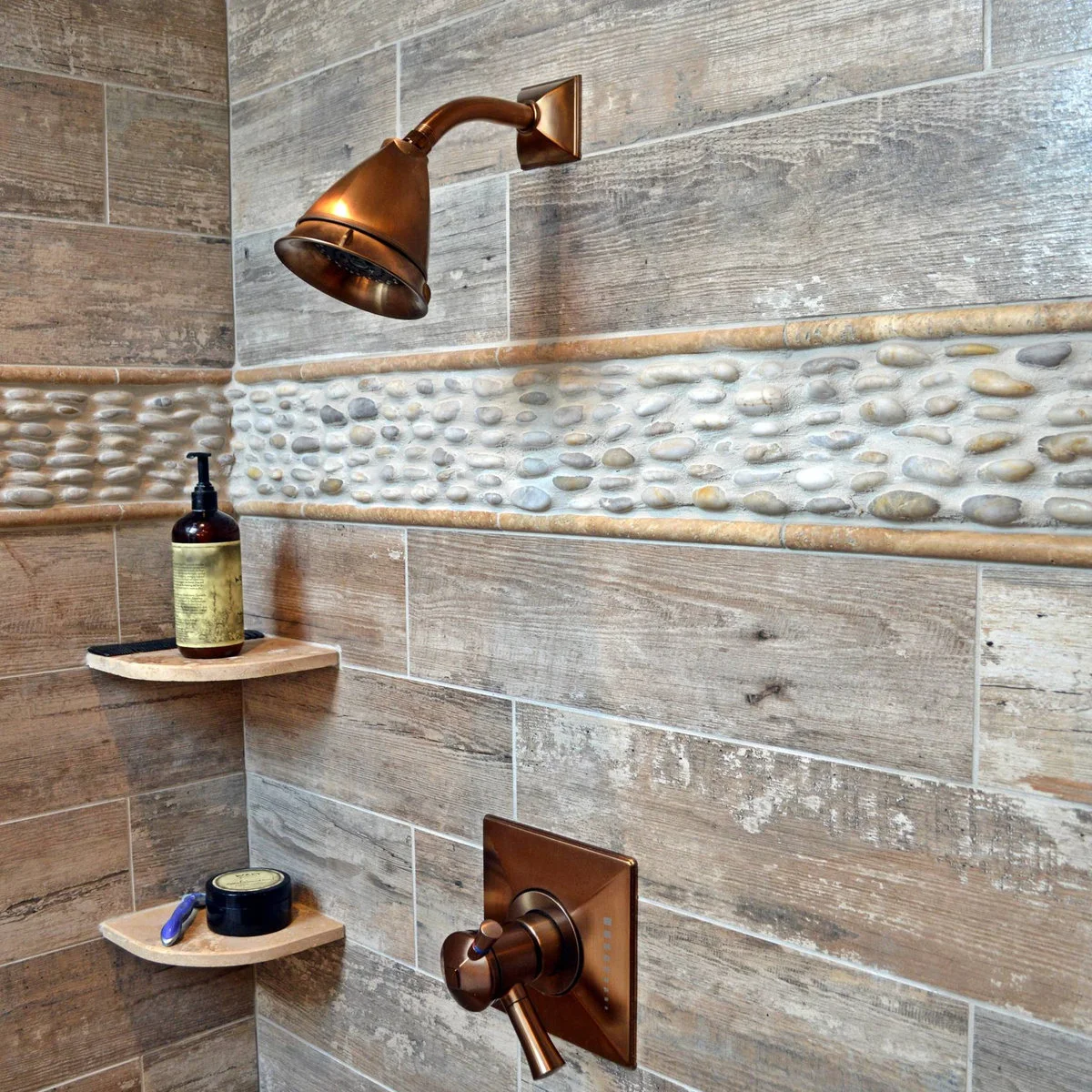 A close view of wood-grain shower tiles with a pebble border, bronze-colored fittings and two small rounded corner shelves.
