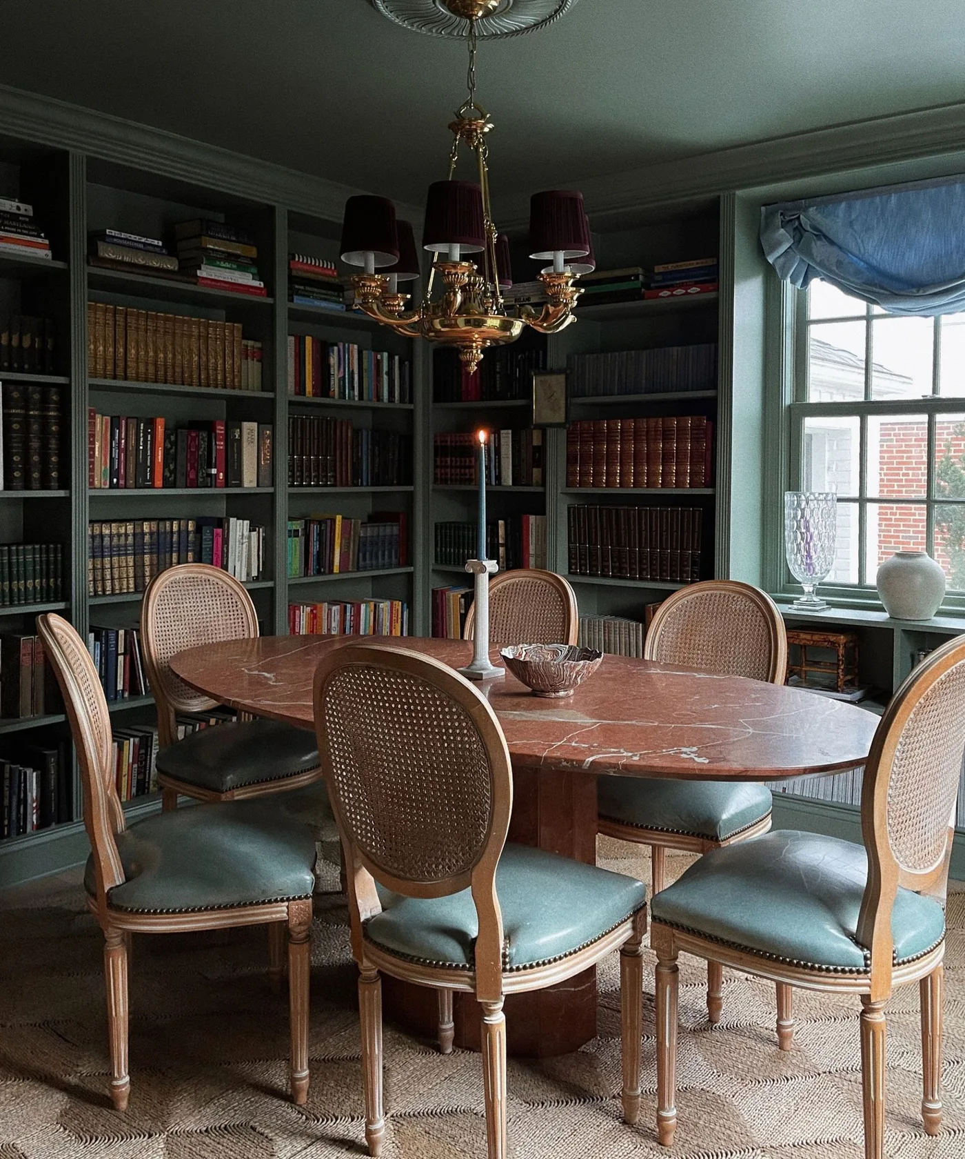 Deep green dining room with perimeter bookshelves, an oval patterned table, cane-backed chairs and a shaded chandelier.