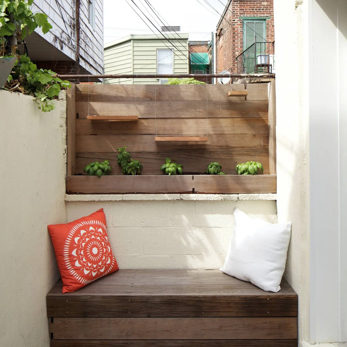 A wood bench with red and white cushions fills a small pale-walled outdoor recess below a wood planter and hanging platforms.