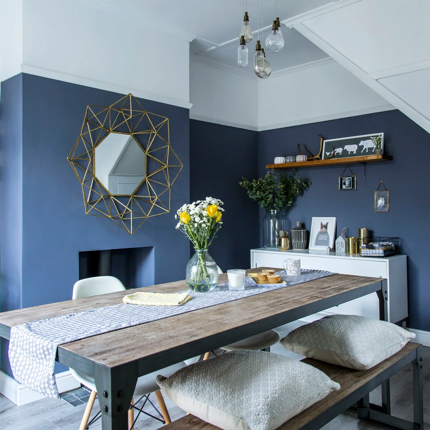 Gold-toned geometric mirror above a fireplace in a navy dining room with a long table and bench