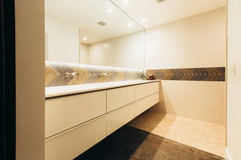 A long floating cream vanity stands below a wide mirror with a narrow dark chevron tile band and wall-mounted taps against pale bathroom walls