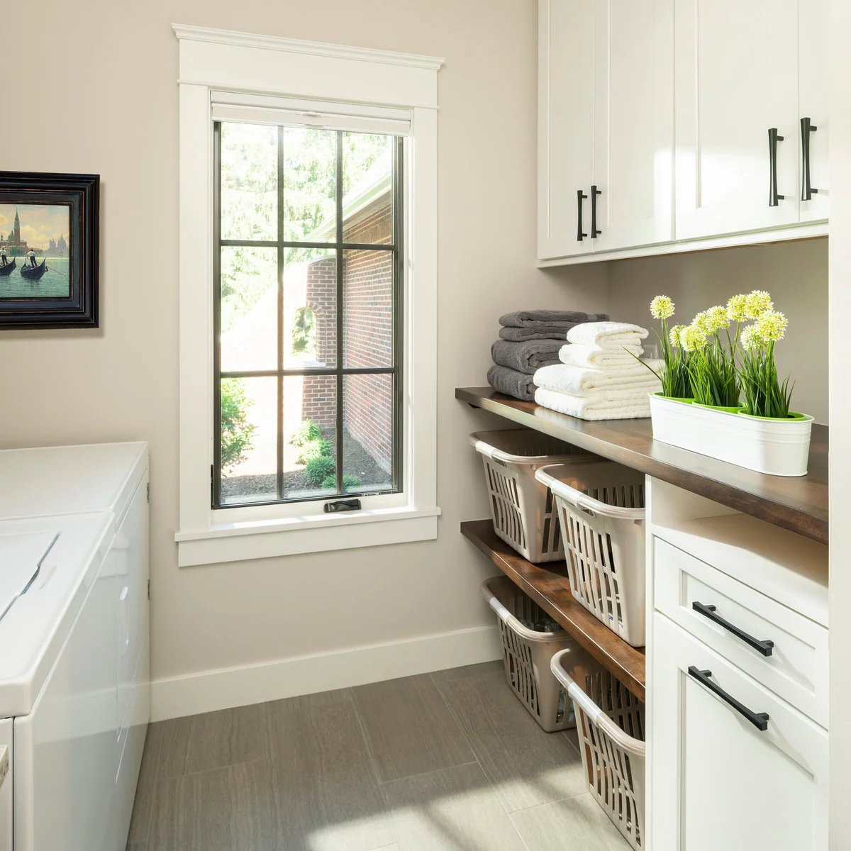 Laundry room with white cabinets, dark open shelving holding four pale baskets and a tall window beside the counter