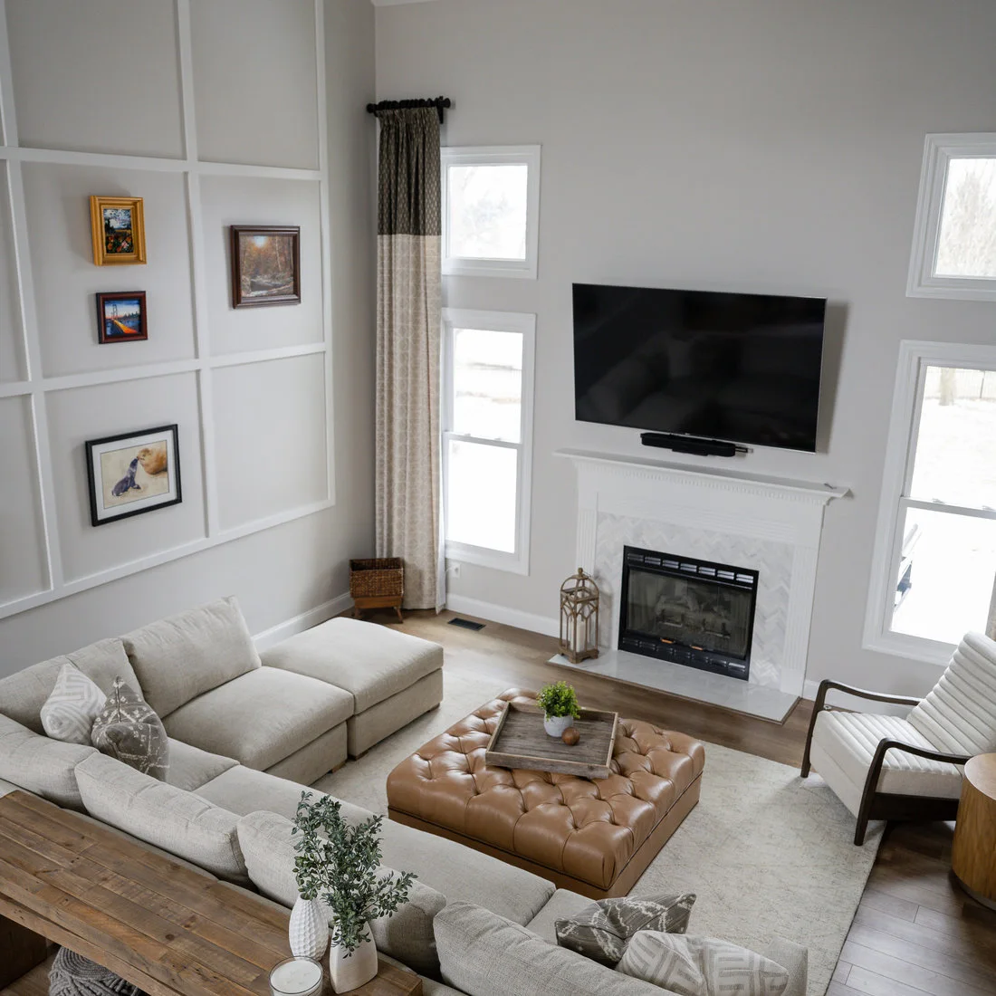 A pale sectional, tan buttoned ottoman and wood-framed chair sit before a white fireplace and light paneled walls.