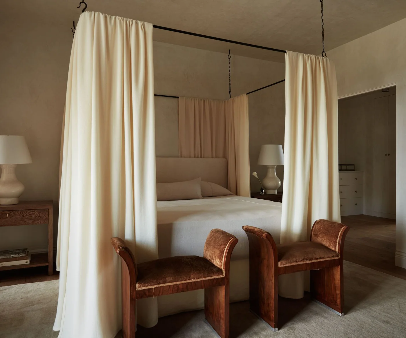 Soft stone-colored bedroom walls and ceiling around a pale curtained canopy bed and brown stools.