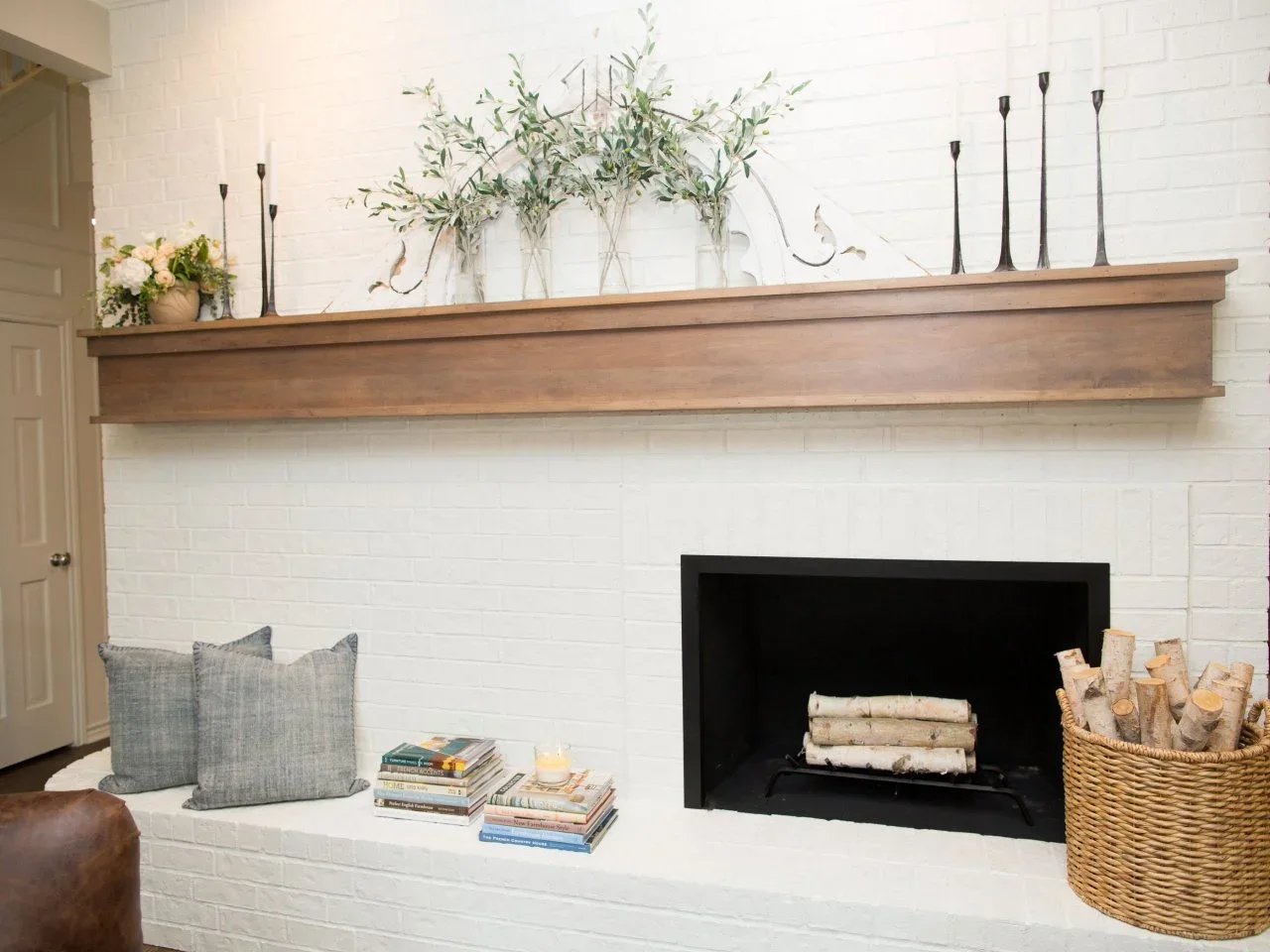 A long timber mantel runs across white-painted brick above a dark fireplace opening and broad hearth.