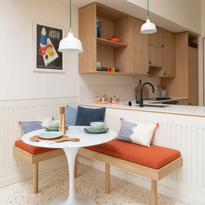 An orange-cushioned wood corner bench surrounds a small white round table beside a light wood kitchen and white paneling.