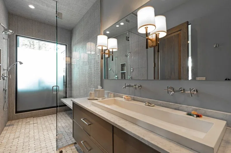 A gray bathroom has a long pale trough-style basin with wall-mounted taps above wood drawers beside glass shower panels and a frosted door
