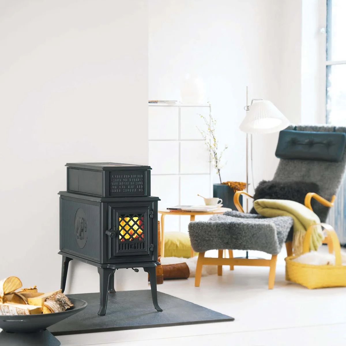 A compact black stove with a patterned glass door stands on a dark rectangular hearth in a pale room beside a gray chair with timber arms.