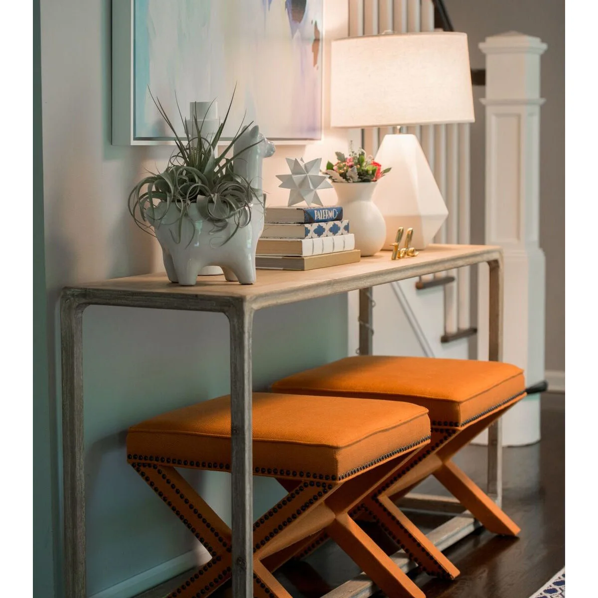 A narrow console with two orange stools beneath it, with a pale lamp, books and a sculptural planter on top.
