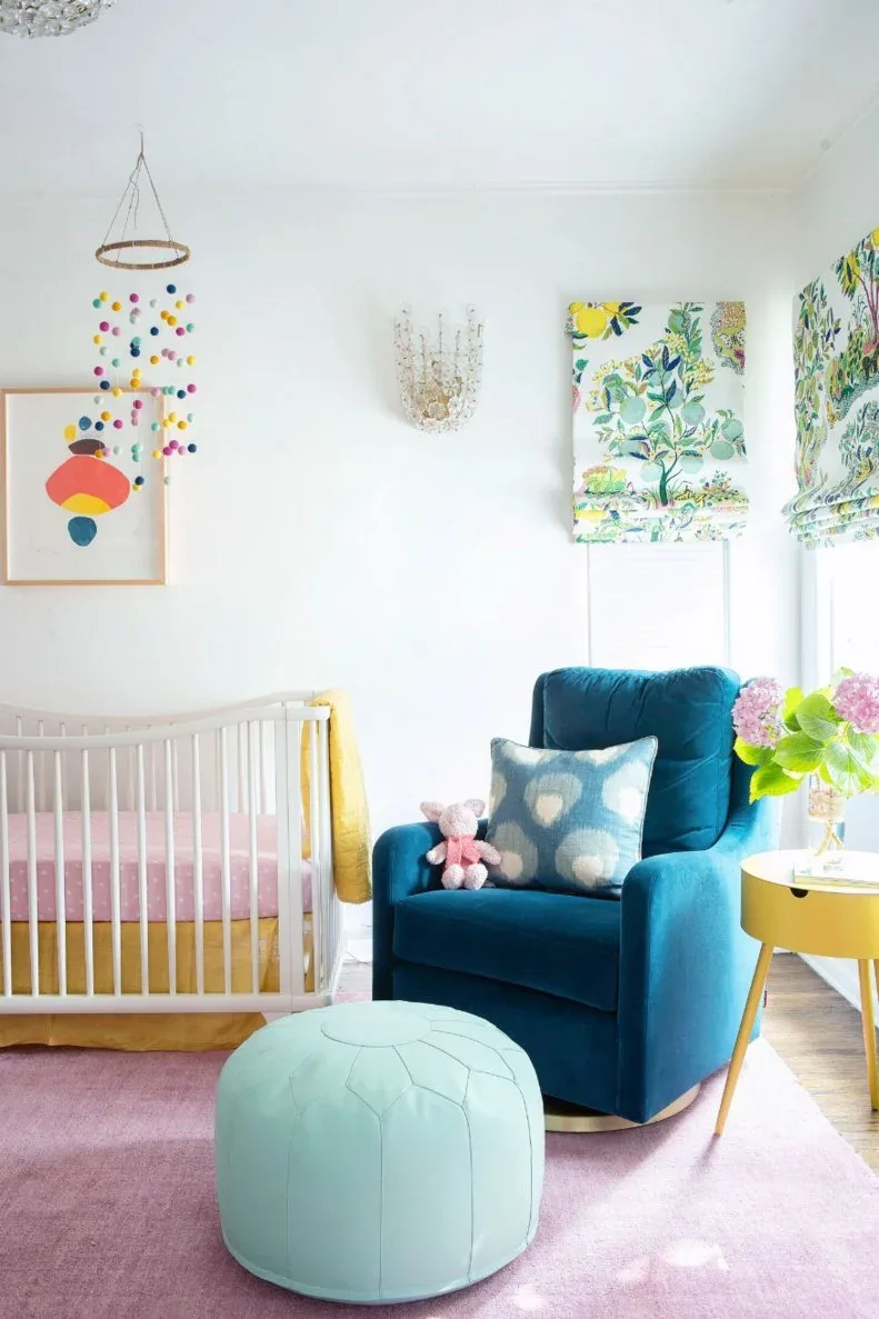 Pale nursery with floral window shades, deep blue adult chair, pale round pouf, yellow table and white crib.