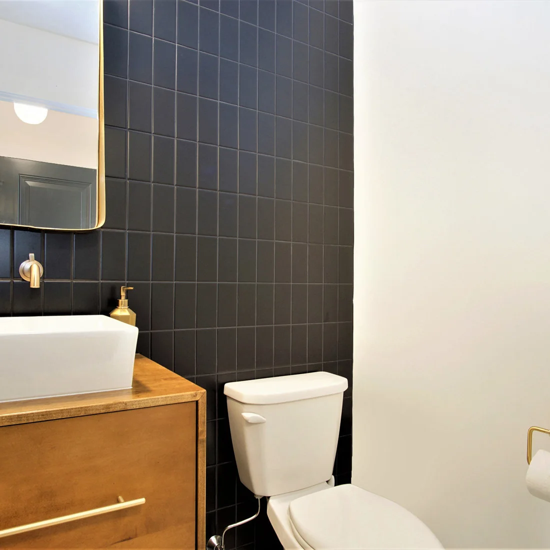 Dark square wall tiles rise behind a wood vanity and white vessel basin, beside a white toilet and plain white side wall with warm-metal fittings.