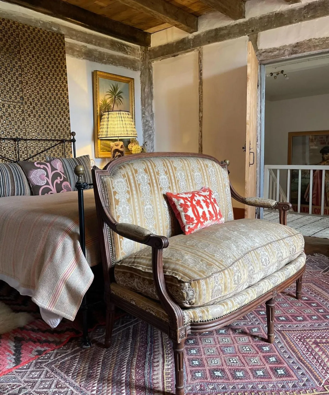 Country bedroom with exposed wall timbers, a patterned upholstered armchair, dark metal bed frame, striped pillows and a small bedside lamp.