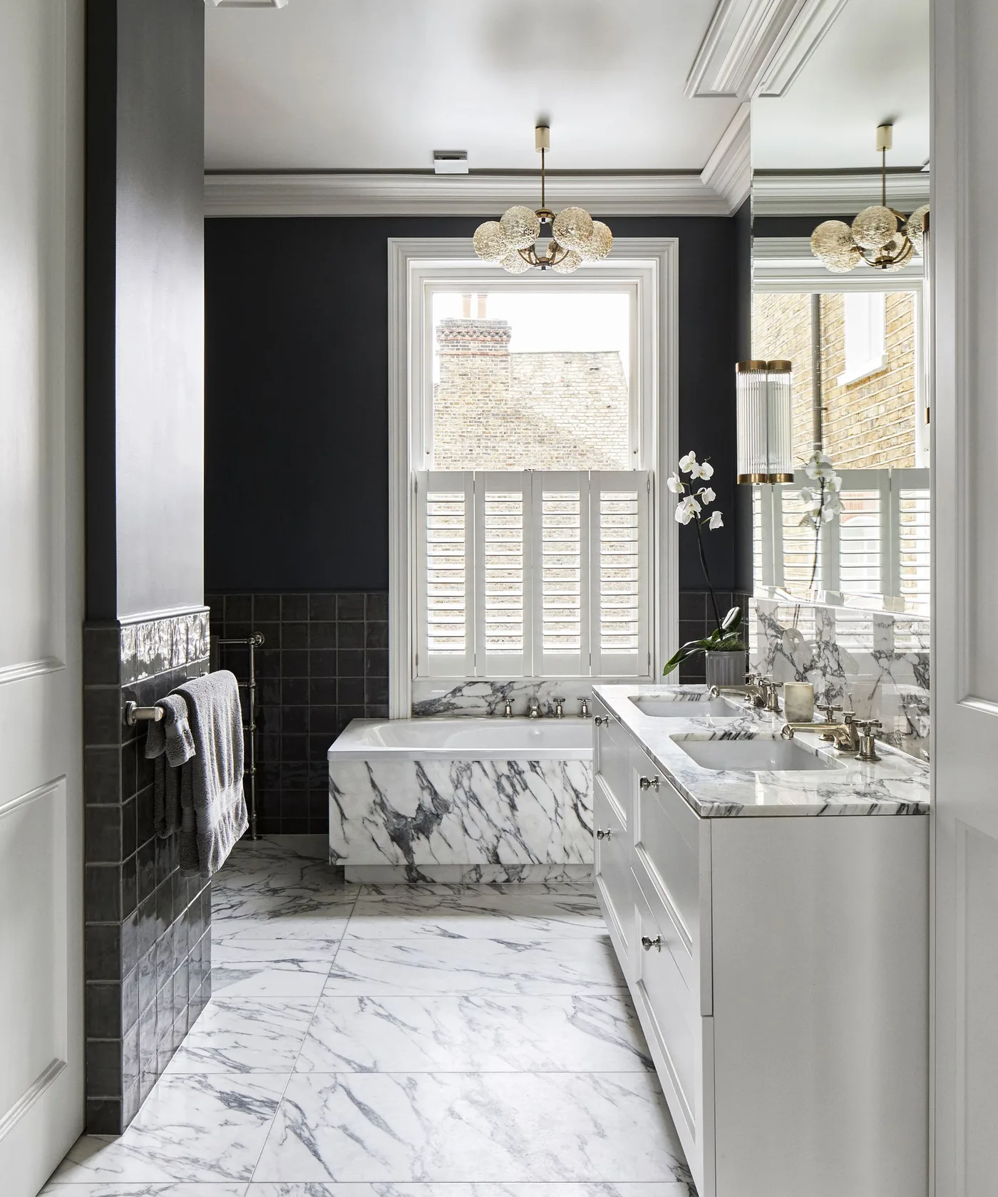 Bathroom with charcoal upper walls, dark square lower tiles, a pale veined bath surround and floor, white vanity, and window shutters.