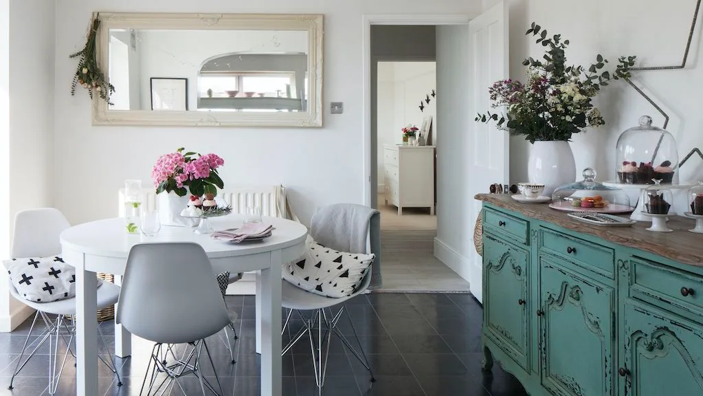 Wide pale rectangular mirror above a small round white table with gray chairs and a turquoise sideboard